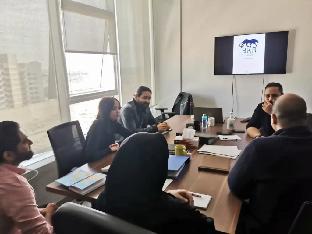 BKR Group staff sitting around a wooden table in an office conversing with the BKR Logo on a screen in the background.